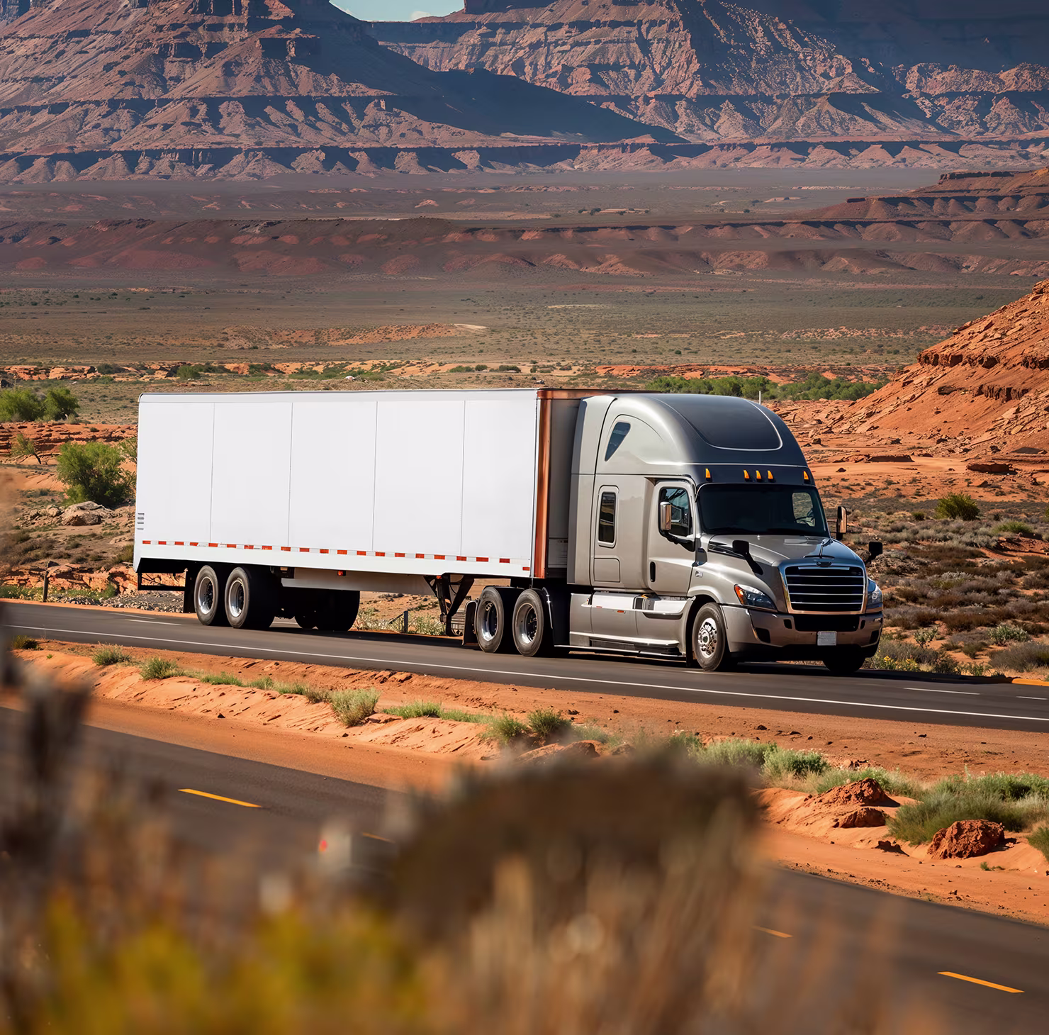 Silver semi truck with a white trailer driving on a highway through a desert landscape with rocky hills.
